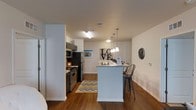 This single-floor residence offers a thoughtfully designed open-concept layout that seamlessly connects the kitchen and living areas, creating a comfortable and functional living environment. The kitchen is a standout feature, equipped with stainless steel appliances — including a gas range, microwave, and refrigerator — complemented by granite-style countertops and dark cabinetry. A peninsula with bar seating serves as both a prep space and casual dining area, illuminated by a trio of elegant pendant lights that add a warm, inviting ambiance. Hardwood-style flooring runs throughout, adding a cohesive and polished look to the space. The living area is generously sized, featuring a ceiling fan with light fixture and a large window that allows natural light to fill the room. The neutral wall palette provides a versatile backdrop, while the well-chosen décor accents contribute to a modern, welcoming atmosphere. A bedroom area is also present, featuring closet storage and clean, neutral finishes that maintain the space's consistent aesthetic. Overall, this single-floor residence balances practicality with contemporary style, making it an attractive and comfortable living space.