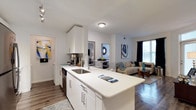 This single-floor residence showcases a thoughtfully designed open-concept layout that seamlessly connects the kitchen and living areas. The kitchen is a standout feature, equipped with crisp white cabinetry, sleek quartz countertops, and a large peninsula with an undermount sink — ideal for both meal preparation and casual dining. Stainless steel appliances, including a dishwasher, add a modern touch, while the peninsula serves as a natural divider between the kitchen and the living space beyond.

The living area benefits from natural light streaming through large windows dressed with dark curtains, creating a bright yet versatile ambiance. Wood-style flooring runs consistently throughout, adding warmth and visual continuity to the space. The open layout provides ample room for a full seating arrangement, with enough space for a sofa, accent chairs, and decorative elements.

A glimpse into an adjacent room reveals a bedroom accessed through a bright doorway, suggesting a functional floor plan. The overall design aesthetic is clean, contemporary, and cohesive, characterized by a neutral white and gray palette accented with colorful wall art.