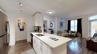 This single-floor residence showcases a thoughtfully designed open-concept layout that seamlessly connects the kitchen and living areas. The kitchen is a standout feature, equipped with crisp white cabinetry, sleek quartz countertops, and a large peninsula with an undermount sink — ideal for both meal preparation and casual dining. Stainless steel appliances, including a dishwasher, add a modern touch, while the peninsula serves as a natural divider between the kitchen and the living space beyond.

The living area benefits from natural light streaming through large windows dressed with dark curtains, creating a bright yet versatile ambiance. Wood-style flooring runs consistently throughout, adding warmth and visual continuity to the space. The open layout provides ample room for a full seating arrangement, with enough space for a sofa, accent chairs, and decorative elements.

A glimpse into an adjacent room reveals a bedroom accessed through a bright doorway, suggesting a functional floor plan. The overall design aesthetic is clean, contemporary, and cohesive, characterized by a neutral white and gray palette accented with colorful wall art.