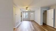 This single-story space is designed with a clean, neutral aesthetic and consistent wood-look flooring that flows throughout each area. The main living space is generously sized, featuring soft gray walls, white baseboards, and a ceiling fan with a light fixture that provides both functionality and a classic touch. Natural light enters through a large sliding glass door fitted with vertical blinds, offering outdoor views of greenery and creating a bright, open atmosphere.

The entryway is welcoming, anchored by a paneled front door with transom glass detail and a nearby closet for convenient storage. A distinctive arched passageway connects the living area to the kitchen, adding an architectural accent. The kitchen features white cabinetry, granite-look countertops, a stainless steel sink, and a dedicated utility room accessible from within.

The bedrooms continue the wood-look flooring and neutral palette, each benefiting from natural light through windows with outdoor views. Closet space is well-distributed, with multiple rooms offering reach-in closets with built-in rods and shelving.

The bathroom is finished in a crisp white scheme, featuring a tub-shower combination, a vanity with storage, and small-format mosaic tile flooring that adds subtle texture to the space.