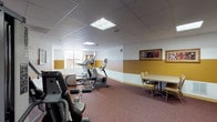 This single-floor fitness room offers a well-rounded exercise environment designed to accommodate a variety of workout needs. The space is decorated with a distinctive two-tone color scheme, featuring a warm gold lower band transitioning to crisp white upper walls, lending the room a welcoming and energetic atmosphere. Durable, multi-toned carpet in earthy red and brown tones covers the floor throughout, providing comfort underfoot during workouts.

The cardio section is well-appointed with multiple Precor elliptical trainers and treadmills arranged along one side of the room, all facing a window that allows natural light to filter in. A multi-station Precor strength-training cable machine occupies a dedicated area, offering users options for resistance and weight training exercises. Framed botanical and nature-themed artwork is displayed on the walls, adding a decorative touch to the functional space.

A small seating and table arrangement in one corner provides a space to rest or review fitness schedules, which are posted on an informational board near the entrance. The room is accessed through elegant wood-framed double doors with glass panel inserts. Recessed panel ceiling tiles with flush fluorescent lighting ensure consistent, even illumination throughout the entire space.