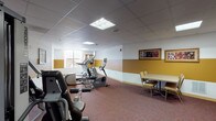 This single-floor fitness room offers a well-rounded exercise environment designed to accommodate a variety of workout needs. The space is decorated with a distinctive two-tone color scheme, featuring a warm gold lower band transitioning to crisp white upper walls, lending the room a welcoming and energetic atmosphere. Durable, multi-toned carpet in earthy red and brown tones covers the floor throughout, providing comfort underfoot during workouts.

The cardio section is well-appointed with multiple Precor elliptical trainers and treadmills arranged along one side of the room, all facing a window that allows natural light to filter in. A multi-station Precor strength-training cable machine occupies a dedicated area, offering users options for resistance and weight training exercises. Framed botanical and nature-themed artwork is displayed on the walls, adding a decorative touch to the functional space.

A small seating and table arrangement in one corner provides a space to rest or review fitness schedules, which are posted on an informational board near the entrance. The room is accessed through elegant wood-framed double doors with glass panel inserts. Recessed panel ceiling tiles with flush fluorescent lighting ensure consistent, even illumination throughout the entire space.