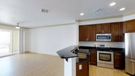 This single-floor residence offers a thoughtfully designed open-concept layout that seamlessly connects the kitchen and living areas, creating a spacious and functional environment. The kitchen is a standout feature, showcasing rich, dark-toned wood cabinetry that provides ample storage, complemented by sleek black granite countertops that extend into a peninsula with an integrated sink — ideal for casual dining or meal preparation. A full suite of stainless steel appliances, including a range, built-in microwave, and dishwasher, lends the kitchen a clean, contemporary aesthetic. Light hardwood flooring runs throughout the space, contributing warmth and continuity between the kitchen and the open living area. The living space is bright and airy, with white walls and recessed lighting that enhance the sense of openness. Crown molding provides a refined architectural detail along the ceiling line, adding a subtle touch of elegance. HVAC vents are neatly integrated into the ceiling, ensuring efficient climate control throughout. The overall ambiance is modern and understated, offering a neutral palette that lends itself well to a variety of interior styles.