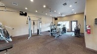 This fitness center is designed to support a comprehensive workout experience within a single, open-plan floor. The space is finished with a distinctive patterned carpet throughout, adding visual interest to the functional layout. Recessed lighting illuminates the room evenly, complemented by natural light streaming in through large windows and a glass-paneled entry door, creating a bright and welcoming environment.

The cardio zone is well-appointed with multiple treadmills and elliptical machines, positioned near the windows to allow exercisers to enjoy outdoor views during their workouts. A wall-mounted flat-screen television is also present for entertainment. The strength training area features a multi-station cable machine and several selectorized resistance machines, offering a range of upper and lower body exercises.

A striking floor-to-ceiling mirrored glass partition with black framing divides a section of the space, enhancing the sense of openness while also providing a dedicated area for stretching or additional activities. The warm, neutral-toned walls contribute to the inviting ambiance, and posted fitness center rules ensure a respectful environment for all users. Security surveillance is noted within the space. Overall, the facility is clean, organized, and thoughtfully laid out to accommodate a variety of fitness routines.