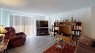 This single-floor residence offers a thoughtfully arranged layout across several distinct living areas. The living room serves as a welcoming central space, featuring light wood-look flooring, a textured ceiling, and a large sliding glass door with vertical blinds that invites natural light and provides access to an outdoor area. The kitchen is a functional highlight, showcasing white shaker-style cabinetry, granite-look countertops, and a full suite of stainless steel appliances including an electric range and refrigerator. The galley-style configuration maximizes efficiency while offering generous counter and storage space. Adjacent to the kitchen, a compact dining area accommodates a glass-top table and provides a programable thermostat for climate control. The primary bedroom features plush carpeting, a window with blackout curtains, and a classic wooden bed frame. A second room, also carpeted, functions flexibly as an additional bedroom or home office, complete with a window for natural light. The full bathroom is finished with wood-look flooring, white cabinetry, granite countertops, a shower-tub combination, and a curved curtain rod. Throughout the space, consistent light flooring and neutral white walls create a clean, cohesive aesthetic.
