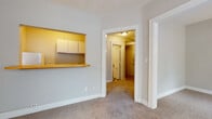 This single-story space is thoughtfully laid out, offering a series of connected rooms with a cohesive neutral color palette throughout. The main living area is generously sized, with plush wall-to-wall carpeting underfoot and multiple windows fitted with horizontal blinds that allow natural light to filter in. A ceiling fan with a built-in light fixture is centered overhead, providing both airflow and ambient illumination. A distinctive architectural feature of this room is the pass-through opening in the wall, framed with warm yellow accents and topped with a wooden ledge countertop, providing a visual and functional connection to the kitchen area beyond. Upper cabinets are visible through the opening, adding storage context to the layout.

A hallway connects the living space to additional rooms, including a secondary carpeted area warmed by a classic cast-iron radiator positioned beneath a window. This room also benefits from natural light and opens to adjacent spaces via a wide, white-trimmed doorway.

The bathroom features a timeless black-and-white checkerboard tile floor, a pedestal sink, a standard toilet, and a mirrored medicine cabinet above — a clean, functional design with a classic character. Overall, the space presents a straightforward and practical layout suited to comfortable everyday living.