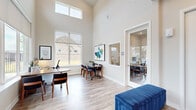 This single-story leasing office is thoughtfully designed to provide a functional and welcoming environment for staff and visitors alike. The main reception area immediately establishes a professional tone, featuring light wood-look flooring, neutral wall tones, and a large wall-mounted display screen alongside a plush blue upholstered bench. A sideboard with decorative accents adds a polished finishing touch to the entryway.

The central open office area is a standout feature, boasting dramatically soaring ceilings accentuated by clerestory windows that flood the space with natural light. Large lower windows frame views of the surrounding exterior, enhancing the sense of openness. Multiple workstations and a small conference table provide ample workspace for staff and client consultations.

A private enclosed office, accessible via a glass-paneled door, offers a quieter setting for one-on-one meetings, equipped with a dedicated desk and seating. Additional consultation seating is available near large windows overlooking the parking area and landscaping.

The kitchenette is well-equipped with white shaker cabinetry, granite countertops, a subway tile backsplash, and a sink, providing a practical break space. Throughout, consistent wood-look flooring and a neutral color palette create a cohesive, professional atmosphere.
