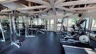 This fitness center combines rustic architectural charm with a comprehensive array of workout equipment. The most striking feature is the soaring timber-frame ceiling, where exposed wooden beams radiate outward in a dramatic pattern, creating an open, airy atmosphere that sets this facility apart from a typical gym environment. Rubber flooring covers the entire workout area, providing both safety and durability underfoot. The space is flooded with natural light through large windows and glass-paneled doors that line the walls, offering unobstructed views of the surrounding wooded landscape. The strength training zone is well-appointed with a power rack, a full dumbbell rack ranging across multiple weight increments, a weight bench, and a medicine ball storage tower stocked with various sizes. Wall-mounted mirrors alongside the strength area enhance the sense of space and allow users to monitor their form. The cardio section features multiple machines, including a treadmill, an elliptical, and additional cardio equipment, all positioned to take advantage of the natural light and outdoor views. A wall-mounted split air conditioning unit ensures climate comfort year-round. The overall ambiance is inviting and motivating, blending natural materials and greenery-framed views with a thoroughly functional fitness environment.