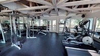 This fitness center combines rustic architectural charm with a comprehensive array of workout equipment. The most striking feature is the soaring timber-frame ceiling, where exposed wooden beams radiate outward in a dramatic pattern, creating an open, airy atmosphere that sets this facility apart from a typical gym environment. Rubber flooring covers the entire workout area, providing both safety and durability underfoot. The space is flooded with natural light through large windows and glass-paneled doors that line the walls, offering unobstructed views of the surrounding wooded landscape. The strength training zone is well-appointed with a power rack, a full dumbbell rack ranging across multiple weight increments, a weight bench, and a medicine ball storage tower stocked with various sizes. Wall-mounted mirrors alongside the strength area enhance the sense of space and allow users to monitor their form. The cardio section features multiple machines, including a treadmill, an elliptical, and additional cardio equipment, all positioned to take advantage of the natural light and outdoor views. A wall-mounted split air conditioning unit ensures climate comfort year-round. The overall ambiance is inviting and motivating, blending natural materials and greenery-framed views with a thoroughly functional fitness environment.