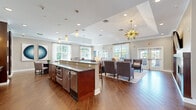 Located at 11 Kensington Ln in Rocky Hill, Connecticut, this single-floor community clubhouse offers a thoughtfully designed, open-concept gathering space that balances elegance with functionality. Rich wood-look plank flooring runs throughout, anchoring the warm, contemporary aesthetic that defines the entire space.

The welcoming entryway corridor features warm-toned accent walls, curated artwork, and a cozy seating nook, setting an inviting tone from the moment you enter. The main gathering area is expansive and light-filled, with large multi-pane windows along multiple walls that frame views of the surrounding greenery. A tray ceiling accented by a striking gold sarburst chandelier crowns the lounge zone, which is furnished with upholstered wingback chairs and accent tables arranged for comfortable socializing.

A central island with granite countertops and bar-height seating serves as a natural hub between the lounge and kitchenette areas. The kitchenette is well-equipped with stainless steel appliances, a coffee and beverage station, a dishwasher, and a utility sink — all finished with granite counters and complemented by modern pendant lighting. A billiards table provides an additional entertainment focal point. French doors lead to an outdoor area, further enhancing the space's connectivity and airflow. The overall ambiance is polished, social, and purposefully designed for community use.