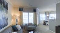 This well-designed single-floor residence offers a comfortable and stylish living environment with a thoughtfully arranged open-concept layout. The living area is anchored by large windows dressed with elegant two-tone cream and teal drapery, allowing abundant natural light to fill the space and create a warm, inviting atmosphere. Crown molding along the ceiling adds a refined architectural touch, complementing the neutral color palette of soft beige walls and plush carpeted flooring.

The living area is generously proportioned, offering ample room for a full seating arrangement, including a sofa and accent chair upholstered in coordinating gray tones, accented with teal and white decorative pillows. A glass-topped coffee table and sleek metallic side accents add a contemporary flair to the space.

Adjacent to the living area, the dining space is seamlessly integrated, featuring a dark wood dining table and chairs, further illuminated by a second set of windows that continue the bright, airy feel throughout. The open sightlines between the two areas enhance the sense of spaciousness and flow.

Overall, this residence presents a clean, sophisticated aesthetic with a cohesive color scheme, quality finishes, and an abundance of natural light that makes the space feel welcoming and move-in ready.