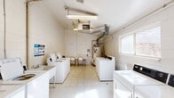 Located at 11501 Dorchester St in Southgate, Michigan, this shared laundry facility is a single-floor space designed to serve the practical needs of its users. The room is constructed with painted concrete block walls and features light-colored tile flooring throughout, giving it a clean and utilitarian appearance.

The facility is well-equipped with multiple top-load washing machines and dryers arranged along both sides of the room, maximizing the available floor space. Utility sinks are also present, providing additional functionality for hand-washing or other laundry tasks. A folding table with chairs is positioned centrally, offering a convenient spot for residents to sort and fold laundry while waiting.

Overhead, fluorescent lighting ensures the space is well-lit, supplemented by natural light from windows on the side and rear walls. A ceiling-mounted HVAC unit and visible ductwork keep the environment comfortable year-round. A water heater with copper piping is also housed within the space. A door at the rear of the room provides direct access to the exterior. Informational signage on the walls offers guidance on proper use of the top-load washers and dryers. Overall, the facility is straightforward and functional, providing essential laundry amenities in a compact, organized layout.