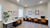 This single-floor business center is thoughtfully designed to support a productive work environment. The main workspace features a long, built-in white countertop workstation along one wall, accommodating multiple users simultaneously, each seat paired with an ergonomic orange rolling chair that adds a vibrant pop of color to the neutral palette. A dedicated computer workstation with a monitor is available for shared use, and a multifunction printer occupies a separate white desk, ensuring essential office tasks can be completed on-site. Natural light filters in through multiple windows dressed with white horizontal blinds, complementing the recessed ceiling lighting that keeps the space bright and functional throughout the day. The floors are finished with a subtle, patterned carpet that adds warmth and texture underfoot. Framed artwork decorates the walls, contributing to a welcoming and professional ambiance. A potted fiddle-leaf fig plant brings a touch of greenery to the space. Adjacent to the main room, a hallway leads to what appear to be private rooms with interior windows and paneled doors, offering additional options for focused work or meetings. The overall design balances practicality with a clean, modern aesthetic.