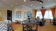 This single-floor space offers a welcoming and versatile lounge environment designed for both casual dining and relaxed socializing. The room features warm hardwood flooring that extends throughout, contributing to an inviting, cohesive aesthetic. A central dining area is anchored by a dark wood table surrounded by classic cross-back chairs with cushioned yellow seats, providing a refined yet comfortable setting for gatherings.

Recessed lighting, complemented by two ceiling fans, ensures the space remains well-lit and comfortable. Crown molding along the ceiling adds a polished architectural detail. The walls are adorned with a curated collection of framed botanical and nature-inspired artwork, enhancing the room's elegant character.

Toward the window-facing side of the room, a separate seating arrangement features upholstered armchairs and accent tables, creating a distinct area for relaxed conversation. Rich, patterned drapery in warm red tones frames the windows, which admit generous natural light while adding depth to the overall design. A flat-screen television is also present, increasing the room's functionality. The overall ambiance is warm, polished, and thoughtfully appointed.