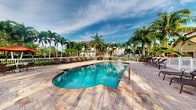 Located at 7575 Hampton Boulevard in North Lauderdale, Florida, this outdoor amenity space showcases a resort-style swimming pool that serves as the centerpiece of the community's recreational offerings. The pool features a freeform shape with clear, turquoise water, complemented by a decorative paver deck that surrounds the entire area. Lounge chairs and seating arrangements are thoughtfully positioned around the pool, providing ample space for relaxation and leisure. Vibrant red patio umbrellas add a pop of color, enhancing the tropical atmosphere. Tall, swaying palm trees frame the space on all sides, creating a lush, private retreat that evokes a quintessential South Florida ambiance. A white perimeter fence encloses the pool area, providing both safety and a clean visual boundary. In the background, a multi-story residential building is visible through the palms, reinforcing the community setting. The open sky above — often filled with the bright Florida sunshine — ensures the space is bathed in natural light throughout the day, making it an inviting outdoor destination year-round.