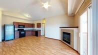 This single-floor space is designed with an open-concept layout that seamlessly connects the living area and kitchen, creating a bright and cohesive environment. The living area is highlighted by soaring vaulted ceilings that amplify the sense of space, complemented by a ceiling fan with integrated lighting. A standout feature is the corner fireplace, elegantly framed with neutral tile surround and a rich wood mantle, serving as a natural focal point for the room. Light hardwood-style flooring extends throughout, adding warmth and continuity across the open floor plan. The kitchen is well-appointed with rich, dark wood cabinetry, dark countertops, and black appliances, including a range and refrigerator, arranged in an efficient L-shaped configuration. A sink is positioned beneath cabinetry along the wall, and a doorway nearby provides convenient access to additional areas of the space. The neutral wall tones throughout create a clean, versatile backdrop that allows for a variety of décor styles. The combination of vaulted ceilings, an open layout, and thoughtful design details makes this space both functional and inviting, suitable for everyday living and entertaining alike.