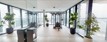 This expansive 5,390-square-foot fitness facility is thoughtfully designed across a single floor, offering a premium workout experience with a sleek, modern aesthetic. Floor-to-ceiling glass walls and oversized windows flood the space with natural light while providing sweeping panoramic views of the surrounding cityscape and waterfront, making every workout visually immersive.

The main workout areas are divided into distinct functional zones. The cardio section features a row of high-end treadmills, stair climbers, ellipticals, and stationary bikes, all positioned to take full advantage of the breathtaking outdoor views. A dedicated strength training area is equipped with multi-functional cable machines, free weights, dumbbells, and adjustable benches, complemented by floor-to-ceiling mirrors that enhance both space and utility.

Exposed concrete columns add an industrial architectural element that contrasts beautifully with the light hardwood flooring and crisp white ceilings. Potted plants are strategically placed throughout, softening the environment and adding a touch of nature. The facility also includes a modern restroom with dark slate tile and ADA-accessible fixtures, as well as a separate lavatory with direct access to an outdoor terrace. Recessed lighting and a clean, uncluttered layout ensure a professional and welcoming atmosphere.