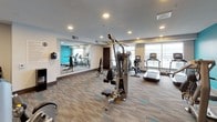 This fitness center is a comprehensively equipped facility designed to support a full range of workout routines. The space is defined by a contemporary aesthetic, highlighted by a bold teal accent wall that contrasts with neutral gray and white tones throughout. Patterned carpet flooring with teal accents adds visual interest underfoot, while recessed lighting provides consistent, even illumination across the entire area.

The cardio zone features multiple treadmills, an elliptical trainer, and a stationary bike, all positioned along large windows that offer outdoor views and allow ample natural light to fill the space. A full-length wall mirror on one side visually expands the room and aids users during workouts.

The strength training area is well-stocked, including a multi-function cable machine, a leg extension/curl machine, an adjustable weight bench, and a dedicated dumbbell rack holding a wide range of free weights. Sanitation stations are positioned conveniently throughout the space. The fitness center is accessed through a single keyed entry door. Posted fitness center rules on the wall reinforce the facility's organized and professional environment.