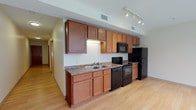 This single-floor unit offers a practical and clean layout designed for comfortable everyday living. The kitchen is a standout feature, equipped with a full suite of black appliances including an electric range, over-the-range microwave, dishwasher, and refrigerator. Warm medium-toned wood cabinetry lines the walls both above and below the counters, providing generous storage space. The laminate countertops offer ample prep area, and a double-basin sink is conveniently positioned along the counter run. Track lighting overhead ensures the kitchen is well-illuminated. Light hardwood-style flooring flows throughout the unit, contributing to a cohesive and airy feel. The living area is brightened by a row of large windows fitted with horizontal blinds, allowing natural light to filter in while maintaining privacy. Low-profile baseboard heaters run along the wall beneath the windows. The bathroom features a full tub and shower combination with chrome fixtures, a toilet, a vanity with a light countertop, and tile flooring. A dropped ceiling with panel tiles adds a defined feel to the bathroom space. Overall, the unit presents a clean, functional environment with consistent finishes throughout.