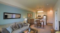 This single-floor space showcases an open-concept layout that seamlessly connects the living, dining, and kitchen areas into one cohesive environment. The living area is anchored by a plush, grey sofa adorned with patterned accent pillows, paired with a dark upholstered ottoman that doubles as a coffee table. A large framed mirror on the sage-green feature wall adds depth and visual interest to the space.

Transitioning naturally toward the dining area, a compact wooden dining table with cross-back chairs provides an intimate setting for meals. The kitchen beyond features stainless steel appliances, including a refrigerator and microwave, set against clean white cabinetry and a dark countertop breakfast bar. A pendant light and a modern track lighting fixture overhead illuminate the kitchen and dining zones warmly.

Throughout the space, soft neutral carpeting grounds the living and dining areas, while the kitchen transitions to a smooth hard floor. Warm, layered lighting creates an inviting ambiance throughout. Framed botanical artwork and carefully placed decorative lamps contribute to the polished, comfortable aesthetic of this well-appointed space.