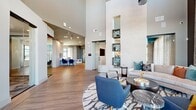 This large-scale amenity facility is designed to impress, offering a comprehensive suite of spaces across a generous single-story footprint. The centerpiece is a soaring double-height lobby and lounge area, where dramatic floor-to-ceiling walls are punctuated by illuminated built-in display niches finished in vibrant teal tile. The space is furnished with contemporary seating arrangements set atop patterned area rugs, complemented by hardwood flooring laid in a herringbone pattern that flows throughout.

An adjacent lounge area features expansive floor-to-ceiling windows framing outdoor views, bathing the room in natural light. A bold large-scale wall mural and a sculptural starburst chandelier serve as striking focal points. A café-style refreshment bar is outfitted with granite countertops, a beverage cooler, microwave, and a geometric dark hexagonal tile backsplash.

The fitness center is well-equipped with strength and cardio equipment, wall-to-wall mirrors, and carpet tile flooring, with direct views to the outdoor pool area. Outside, a covered patio with turf putting green connects to the pool courtyard. The exterior features limestone and board-and-batten siding with covered walkways, offering a polished, resort-style atmosphere throughout.