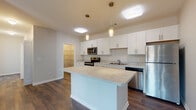 This single-floor residence is designed with a clean, contemporary aesthetic that emphasizes functionality and an open layout. The kitchen serves as a central highlight, featuring white shaker-style cabinetry extending to the upper walls, light stone-look countertops, and a full suite of stainless steel appliances including a range with over-the-range microwave, dishwasher, and refrigerator. A spacious center island with matching countertop provides additional workspace and casual seating. Pendant lighting with brass accents adds a warm, modern touch overhead, while a walk-in pantry offers generous storage. Dark hardwood-style flooring flows seamlessly through the kitchen and open living area, creating a cohesive and inviting atmosphere.

The bedrooms are carpeted in neutral tones, offering comfort underfoot, and each is naturally lit through slatted windows. The primary bedroom includes a ceiling fan and access to an en-suite bathroom featuring white vanity cabinetry with stone-look countertops. A secondary full bathroom includes a tiled tub-shower surround in a warm wood-look tile pattern. The neutral color palette throughout—soft whites, warm beiges, and earthy tones—gives the entire space a bright, move-in-ready feel.