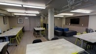 This single-floor communal space is thoughtfully designed to accommodate a range of activities, from collaborative work sessions to casual socializing. The open-plan layout is anchored by structural columns that subtly divide the area into distinct functional zones without sacrificing the sense of openness throughout.

The dining and work area features a large rectangular table surrounded by a mix of wheeled task chairs and vibrant yellow seating, offering a dynamic environment for group work or shared meals. Light wood-toned flooring runs throughout, lending warmth to the otherwise neutral interior palette of grey and taupe walls.

A dedicated lounge zone is furnished with a generously sized modular sectional sofa in grey and blue upholstery, positioned to face a wall-mounted flat-screen television, creating an inviting space for relaxation or group viewing. Adjacent to the lounge, a built-in cabinetry unit with a countertop provides additional surface space, likely serving as a refreshment or utility station.

Overhead, exposed ductwork and fluorescent linear lighting fixtures illuminate the space evenly, contributing to its functional, modern aesthetic. Additional seating along the perimeter, including a row of yellow stools at a counter, enhances the space's flexibility. Overall, the layout reflects a well-considered design suited for community engagement and multipurpose use.