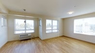 This single-floor space offers a clean, functional layout highlighted by an abundance of natural light streaming through multiple large windows. The windows frame views of surrounding trees and the exterior environment, creating a connection to the outdoors while keeping the interior bright and airy throughout the day.

The main living area is finished with light-toned hardwood flooring that runs throughout, contributing to a warm yet minimalist aesthetic. The neutral, cream-colored walls complement the flooring and enhance the sense of openness within the space. A wall-mounted HVAC unit is positioned beneath one of the primary windows, providing efficient climate control for year-round comfort.

Ceiling-mounted and pendant lighting fixtures supplement the natural light, ensuring the space remains well-lit during evening hours. The smooth white ceiling and clean architectural lines give the interior a straightforward, uncluttered feel. The overall ambiance is calm and practical, with a move-in-ready character suited to a variety of uses. The layout is open and easily adaptable, making it a versatile and functional living space.