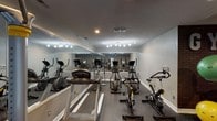 Located at 5803 W 10th St in Indianapolis, this single-floor fitness space is designed to function as a comprehensive private gym. The room is finished with durable rubber flooring throughout, providing a practical and safe surface suited for a wide range of exercise activities. Neutral gray walls and white baseboards give the space a clean, polished appearance, while a textured ceiling houses recessed lighting and track lighting that ensure the area is well-illuminated.

A defining feature of the space is the large floor-to-ceiling mirror spanning one full wall, which visually expands the room and allows users to monitor form during workouts. The cardio section is well-stocked with multiple elliptical trainers, stationary spin bikes, and a treadmill. A dedicated strength-training area houses a Hoist cable machine, a weight bench, and a rack of dumbbells. An accent wall featuring exposed brick and a bold "GYM" sign adds character, and a storage rack holds stability balls, medicine balls, and resistance accessories. Louvered double doors provide additional storage. Natural light enters through a window, and a ceiling fan aids in ventilation, contributing to a functional and comfortable workout environment.