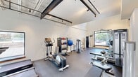 Located at 1409 West Broad Street in Quakertown, PA, this fitness facility offers a thoughtfully designed exercise space that balances function with modern aesthetics. The single-floor layout provides a generous open floor plan, accommodating a wide variety of workout equipment without feeling cramped.

The exercise room features soaring ceilings that lend a sense of openness and airiness, complemented by an abundance of natural light streaming through large windows that offer views of the surrounding greenery. Industrial-style exposed steel beams and track lighting run overhead, adding a sleek, contemporary character to the space.

The fitness area is comprehensively equipped with a full range of Life Fitness strength machines, free weights, a dumbbell rack, cable machines, a squat rack with bench, and cardio equipment including treadmills, an elliptical, and a stationary bike. Stability balls and kettlebells round out the functional training options. The durable gray flooring, including padded mat sections, provides a safe and practical surface throughout.

A striking floor-to-ceiling steel-framed glass partition wall with a wide doorway connects the exercise room to an adjacent corridor, allowing natural light to flow freely while maintaining a sense of defined space. The overall ambiance is clean, bright, and professional, making this facility well-suited for a full range of fitness activities.