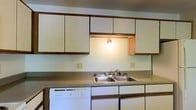 This single-floor residence offers a straightforward and functional layout designed to make the most of its space. The kitchen is a central feature of the home, showcasing a clean, utilitarian design with an abundance of white upper and lower cabinetry trimmed in warm wood-toned edging, providing generous storage throughout. A continuous countertop runs the length of the kitchen, offering ample workspace for meal preparation. The double-basin stainless steel sink sits beneath a pass-through opening, which allows natural interaction between the kitchen and the adjoining area while also letting in additional light. An under-cabinet light fixture provides warm, focused illumination over the sink area, adding a welcoming glow. A built-in dishwasher is seamlessly integrated beneath the counter, contributing to the kitchen's practical appeal. The light-toned walls and neutral surfaces lend the space a clean, open feel. Overall, this residence presents a no-frills, well-organized living environment that prioritizes function and ease of use, making it a practical choice for those seeking a comfortable and efficient single-level home.