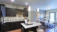 This single-floor space offers a thoughtfully designed open-concept layout that seamlessly connects the kitchen and living areas, creating a warm and inviting atmosphere. The kitchen is a standout feature, showcasing rich dark cabinetry with sleek hardware, light granite countertops, and a central island that doubles as a casual dining and entertaining hub. Pendant lighting hangs above the island, adding a modern touch, while recessed ceiling lights provide ample illumination throughout. A double-basin sink and full appliance suite complete the kitchen's functional appeal. The open flow into the living area creates a sense of spaciousness, with the two zones working in harmony for everyday living and hosting. Neutral wall tones in soft beige and gray complement the dark cabinetry and light countertops, striking a contemporary balance. Hardwood-style flooring runs throughout, adding warmth and continuity to the space. The living area is bright and airy, benefiting from natural light and a comfortable, modern furnishing arrangement. Overall, this space combines practical design with stylish details, making it a welcoming and highly functional single-level home.