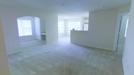 This single-floor space offers an open-concept layout that seamlessly connects the living area to the kitchen, creating a sense of spaciousness and ease of flow. The living area is finished with light-toned wall-to-wall carpeting that extends throughout, providing a clean and neutral foundation. Large windows along multiple walls allow generous amounts of natural light to fill the interior, contributing to an airy, bright atmosphere.

The kitchen is partially separated from the living area by a raised breakfast bar or peninsula, which serves as a natural visual divider while maintaining the open feel. From this vantage point, white cabinetry and a refrigerator are visible within the kitchen, suggesting a functional and simply appointed cooking space. The countertops appear light in color, consistent with the overall neutral palette of the unit.

To one side of the living area, an arched doorway leads to an additional room, partially visible and appearing to contain built-in shelving or cabinetry. The walls throughout are painted in a soft, warm white tone, and the baseboards are cleanly finished. Overall, the space presents a well-lit, move-in-ready interior with a cohesive and understated aesthetic.