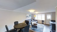 This single-floor space offers a seamlessly integrated living and dining area designed with comfort and functionality in mind. The open-plan layout creates a generous sense of space, allowing natural light to flow freely through large windows that brighten the entire room.

The living area is anchored by a plush gray sofa and matching armchair, arranged around a sleek coffee table atop a patterned blue and white area rug. A stylish floor lamp complements the flush-mount ceiling light fixtures, providing layered illumination throughout the space. Two framed abstract artworks in soft watercolor tones add a touch of personality to the neutral walls.

The dining area features a classic round dark-wood table paired with tufted upholstered chairs, creating an intimate setting ideal for meals or casual gatherings. The clean white ceiling and warm greige walls establish a cohesive, modern aesthetic throughout.

Overall, the space exudes a calm and polished ambiance, combining contemporary furnishings with a restrained color palette of grays, blues, and warm neutrals. The thoughtful arrangement of furniture and décor makes efficient use of the available space while maintaining an open, airy feel that is both functional and inviting.
