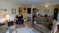 This single-floor residence offers a thoughtfully arranged living space with a warm, comfortable ambiance throughout. The living room serves as the heart of the home, featuring plush carpeting, a cushioned sofa and recliner armchair, and a decorative area rug that anchors the seating arrangement. A classic wooden display cabinet with glass doors adds a traditional touch, providing an elegant space for keepsakes and collectibles. Framed artwork adorns the walls, contributing to the homey character of the room. A ceiling fan overhead provides both comfort and light. Adjacent to the living area, a dining space is visible, furnished with a wooden table and chairs, suitable for everyday meals and gatherings. The open sightline into the kitchen reveals wood-toned cabinetry and a refrigerator, indicating a functional and accessible layout. A small side table with a decorative lamp adds a charming accent near the seating area. The neutral, light-toned walls and carpeting throughout create a calm, cohesive aesthetic, while the open flow between the living, dining, and kitchen areas makes the space feel connected and easy to navigate.
