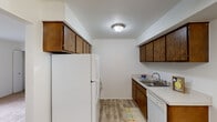 This single-floor unit is organized with a practical, efficient layout that connects key living areas seamlessly. The galley-style kitchen is lined on both sides with dark wood cabinetry, providing ample upper and lower storage. White appliances, including a full-sized refrigerator and dishwasher, complement the light countertops, while wood-look plank flooring adds warmth underfoot and a flush-mount ceiling fixture provides overhead lighting.

The living area is a well-proportioned, open space finished with neutral carpet and bright white walls. A large window fitted with vertical blinds allows natural light to fill the room, contributing to an airy atmosphere. An adjacent carpeted room, visible through a connecting doorway, opens toward both the kitchen and a hallway, offering flexible use of space. A bifold closet door provides additional storage in this area.

The bedrooms are carpeted and feature white walls, with multiple windows dressed in vertical blinds that invite natural light. One bedroom includes a walk-in closet alongside a standard closet, offering generous storage capacity.

The bathroom is compact and functional, featuring wood-look flooring, a tub-shower combination, a toilet, and a vanity with storage. Overall, the unit maintains a clean, neutral palette throughout.