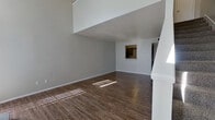 This two-story space is efficiently designed across two floors, with the larger ground floor housing the primary living areas and the upper level providing additional functional space. The combined kitchen and living room on the ground floor serves as the heart of the home, featuring durable wood-style flooring that runs throughout. The living area is particularly striking, boasting dramatically high ceilings that create an open, airy atmosphere, flanked by tall vertical-blind windows that allow abundant natural light to pour in. A tiled fireplace with a raised hearth serves as a focal point in this space, adding character and warmth.

The kitchen is efficiently laid out, featuring gray cabinetry, speckled countertops, a stainless steel sink, a dishwasher, and a full-size black refrigerator. A pass-through window connects the kitchen to the living area, enhancing the open feel between the two spaces. The ground floor also includes a hallway leading to one of the two bathrooms, which is equipped with a combined tub and shower, a speckled vanity countertop, gray cabinetry, and a large mirror. The upper level, accessed via a staircase, contains the second bathroom, which includes a towel bar and toilet.