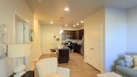 This single-floor space showcases a thoughtfully designed open-concept layout that integrates the living and kitchen areas into one cohesive environment. Light hardwood flooring runs throughout, contributing to a bright and airy atmosphere complemented by warm recessed lighting across the ceiling.

The kitchen is a standout feature, outfitted with dark espresso cabinetry that contrasts elegantly against the light walls and floors. Stainless steel appliances, including a refrigerator, range, and built-in microwave, provide a modern and functional aesthetic. A central kitchen island with a dark countertop offers additional workspace and seating with bar-height stools, making it ideal for casual dining or entertaining. Pendant lights hang above the island, adding a refined finishing touch.

The living area flows naturally from the kitchen, with enough room to accommodate comfortable seating. White-paneled doors provide access to additional rooms, maintaining a clean and consistent design language. The overall ambiance is modern and welcoming, with a neutral color palette that enhances the sense of openness throughout the space.