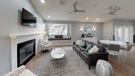 This thoughtfully designed single-floor space offers an open, flowing layout united by warm hardwood-style flooring and soft grey walls accented with two-tone wainscoting details. Vaulted ceilings with recessed lighting and ceiling fans lend a sense of airiness throughout, while large windows and French doors flood the interior with natural light and offer views of surrounding greenery.

The main lounge area is anchored by a white-surround fireplace with a wall-mounted television above, complemented by a built-in window bench seat dressed in teal and grey cushions. A dark leather sofa, patterned accent chairs, and a round white coffee table complete the seating arrangement atop a textured area rug. Adjacent to the lounge, a secondary seating nook features cream upholstered chairs paired with teal-accented pillows and framed artwork overhead.

A standout feature is the rustic wood-paneled bar counter with a white quartz top and modern grey barstools, functioning as a natural divider between the lounge and a separate dining area with a round white table. A beverage station and decorative mirror wall sconces add functional elegance. French doors lead to an additional room, enhancing the versatility of this well-appointed space.