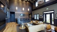This single-floor space combines the ambiance of an upscale lounge with the functionality of a social gathering venue. The most striking architectural feature is the dramatically vaulted ceiling, accented by dark exposed beams that draw the eye upward and create an impressive sense of height and grandeur. A combination of pendant lighting and a statement chandelier illuminates the space warmly throughout.

The bar area serves as a central focal point, featuring a tiled backsplash with a diamond-pattern design, a neatly arranged display of wine and spirit bottles along the back wall, and a mounted flat-screen display. A row of dark bar stools lines the counter, making it ideal for casual seating.

Adjacent to the bar, a comfortable lounge area is furnished with tan leather sofas and blue upholstered accent chairs arranged around a low coffee table, creating an inviting space for conversation. Light hardwood flooring runs throughout, providing warmth against the darker wall tones and trim. Large windows with dark frames allow natural light to filter in, offering views of the surrounding greenery and enhancing the overall welcoming atmosphere of the space.