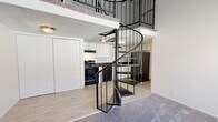 This residence offers a distinctive layout highlighted by a wrought-iron spiral staircase that serves as the space's central architectural feature, connecting the main living level to an upper loft area. The main level features light wood-style flooring flowing into a functional kitchen equipped with white cabinetry, dark speckled countertops, a double stainless steel sink, and black appliances including a gas range and dishwasher. A closet with bifold doors provides additional storage near the kitchen area. A full bathroom on the main level includes a tiled shower-tub combination, dark countertop vanity, and wood-style flooring. The upper loft level, accessed via the spiral staircase, features vaulted ceilings with a textured finish and a ceiling fan, along with carpeted flooring, a built-in closet, and an iron railing overlooking the level below. Bedrooms are carpeted and bright, each featuring vaulted or angled ceilings, closets, and windows that bring in natural light. A second bathroom on this level includes a standalone shower with frosted glass door and a dark countertop vanity. Throughout the space, neutral tones and clean lines create a calm, functional ambiance.