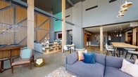 Located at 195 E Main St in the heart of Columbus, Ohio, this single-floor space delivers an impressive blend of industrial architecture and contemporary design. The centerpiece of the space is a dramatic double-height main lounge area, defined by soaring ceilings, exposed structural columns clad in warm wood, and a sweeping wall of floor-to-ceiling windows that flood the interior with natural light while framing panoramic views of the downtown Columbus skyline. Polished concrete floors run throughout, grounding the space with an urban, loft-like character.

The open layout accommodates multiple distinct seating areas, including casual lounge zones with modern sofas and accent chairs, and a more intimate sitting nook anchored by a bold wood-and-steel feature wall adorned with geometric paneling and subtle lighting. A glass-top round table set atop a zebra-print rug serves as a stylish meeting or gathering spot, complemented by living moss wall accents and pendant lighting nearby.

A well-appointed beverage station features a Starbucks coffee machine, white cabinetry, a stainless steel under-counter refrigerator, and a vibrant teal hexagonal tile backsplash with floating wood shelves. Together, these elements create a dynamic, hospitality-forward environment well-suited for collaboration, community, or professional use.