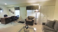 This two-floor space offers a practical and comfortable layout designed for everyday living. The main living area sets a welcoming tone with its neutral, light-toned walls and carpeted flooring that adds warmth underfoot. A plush armchair and sofa are arranged around a glass-topped coffee table, creating an inviting seating arrangement ideal for relaxing or receiving guests. A classic floor lamp provides additional ambient lighting, complementing the natural light that filters in through multiple windows dressed with dark valances. The entryway is clearly defined, featuring a solid front door framed by a decorative fabric valance, with a welcome mat grounding the transition between indoors and out. A second door nearby offers additional access or storage. The space also includes a dedicated seating nook to one side, furnished with dark chairs, which could serve as a reading corner or small workspace. The overall ambiance is understated and functional, with a cohesive neutral color palette running throughout. Spanning two floors, the property provides ample room for varied uses, with the ground level establishing a calm, lived-in atmosphere that balances comfort and practicality.