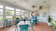This single-floor community space combines a welcoming dining area and a functional kitchen in a compact yet efficiently arranged layout. Throughout the space, distinctive herringbone-patterned brick flooring anchors the design, adding warmth and texture underfoot.

The main gathering area is bathed in natural light, courtesy of expansive white-framed windows that line the walls and offer views of the surrounding greenery and community grounds. White shiplap walls and a tongue-and-groove ceiling contribute to a clean, coastal aesthetic, while a ceiling fan ensures comfortable airflow. Modern white dining tables paired with vibrant teal chairs create a lively, inviting atmosphere suited for socializing and casual gatherings. A wall-mounted television adds a practical amenity to the space.

The kitchen is thoughtfully appointed with white cabinetry, a tile backsplash, light countertops, and stainless steel appliances, including a microwave and a professional-grade range with a large ventilation hood. A built-in grill station with an overhead exhaust hood serves as a standout feature, making the kitchen well-suited for group events and catered functions. A sink and ample counter space round out the kitchen's practical offerings.

Together, the two areas form a versatile, cheerful space that balances functionality with an airy, resort-inspired design sensibility.