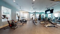 Located at 2600 Executive Drive in Venice, Florida, this single-floor fitness center is a well-appointed facility designed to support a comprehensive workout experience. The space is thoughtfully organized into distinct functional zones, allowing members to seamlessly transition between cardio, strength training, and group exercise activities.

The cardio area is stocked with spin bikes, elliptical machines, treadmills, and rowing machines, all positioned near large windows that allow natural light to fill the space. Ceiling fans throughout ensure comfortable airflow during workouts. A dedicated cycling zone features a flat-screen television mounted against a striking decorative wall accent of angled wooden slats, creating an energizing atmosphere.

The strength and free weights section is anchored by a full rack of dumbbells, multiple adjustable benches, and a multi-station cable machine. Floor-to-ceiling mirrors line the walls, visually expanding the room and allowing users to monitor their form. A ceiling-mounted television provides entertainment during training sessions.

A convenient amenity nook near the entrance includes a compact refrigerator and wall-mounted shelving stocked with yoga mats, catering to flexibility and wellness routines. The overall design palette of soft teal and neutral tones, combined with durable rubberized flooring, gives the facility a clean, modern, and professional ambiance suited for all fitness levels.