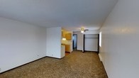 This single-floor unit features an open-concept layout that combines the living and sleeping areas into one cohesive space. The main area is finished with wall-to-wall carpet in a neutral brown tone, providing a warm and practical foundation throughout. White walls keep the interior bright and versatile, complemented by dark baseboards that add a subtle contrasting detail.

A ceiling fan with a light fixture is centrally positioned overhead, offering both air circulation and ambient lighting to the space. Along one wall, a wide closet with a built-in hanging rod and shelf provides convenient storage directly within the main living area.

The kitchen is partially separated by a short partition wall, creating a defined cooking zone without fully closing off the space. It features honey-toned wood cabinetry, an under-cabinet light, and a sink, with room for standard appliances. A doorway from the kitchen leads to an additional area, likely a bathroom or utility space.

The textured ceiling adds a classic finishing detail, and the overall layout is straightforward and functional, making efficient use of the available footprint.