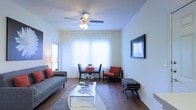 This single-floor residence offers a thoughtfully designed open-concept layout that seamlessly connects the main living spaces. The living room is bright and welcoming, featuring warm hardwood-style flooring, a ceiling fan with integrated lighting, and large windows dressed with sheer white curtains that fill the room with natural light. A wall-mounted flat-screen TV and a glass-top coffee table complement the space, while a cozy sofa and accent chair with bold red cushions add a pop of color and warmth.

Adjacent to the living room, a compact dining nook accommodates a round table and chairs, making efficient use of the space near the windows. The open sightlines into the kitchen reveal dark espresso cabinetry paired with light countertops, creating a striking contrast. The kitchen is well-equipped and functional, featuring ample cabinet storage and under-cabinet lighting. A breakfast bar with stool seating bridges the kitchen and the main living area, enhancing the open flow of the floor plan.

Consistent hardwood-style flooring runs throughout, tying the spaces together cohesively. Clean white walls and neutral tones create a bright, airy ambiance, while warm accent colors and modern furnishings give the space a polished, contemporary feel throughout.