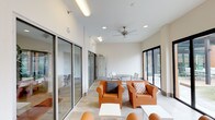This well-appointed single-floor space serves as a stylish communal lounge and gathering area, defined by its clean, contemporary architecture and thoughtfully curated furnishings. The interior is anchored by crisp white walls and smooth tile flooring that extend throughout, creating a cohesive and airy atmosphere. Large black-framed floor-to-ceiling sliding glass doors and glass panel walls line the perimeter, flooding the space with abundant natural light and offering unobstructed views of the surrounding outdoor courtyard and urban skyline beyond.

The lounge area features a grouping of bold orange club chairs accented with decorative throw pillows, arranged around low white side tables to create an inviting conversation setting. A ceiling fan overhead adds a functional touch while contributing to the room's relaxed ambiance. A dining area with a round table and metal chairs provides an additional gathering spot, ideal for casual meetings or meals.

A compact beverage station with a Keurig coffee maker and ample storage sits conveniently along one wall, adding everyday practicality to the space. Glass partitions offer views into adjacent interior rooms, including a warmly lit lounge with lush hanging plants and a fireplace. Outside, a furnished patio with matching orange seating, a pergola structure, and a fireplace extends the living space seamlessly into the open air.