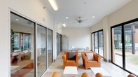 This well-appointed single-floor space serves as a stylish communal lounge and gathering area, defined by its clean, contemporary architecture and thoughtfully curated furnishings. The interior is anchored by crisp white walls and smooth tile flooring that extend throughout, creating a cohesive and airy atmosphere. Large black-framed floor-to-ceiling sliding glass doors and glass panel walls line the perimeter, flooding the space with abundant natural light and offering unobstructed views of the surrounding outdoor courtyard and urban skyline beyond.

The lounge area features a grouping of bold orange club chairs accented with decorative throw pillows, arranged around low white side tables to create an inviting conversation setting. A ceiling fan overhead adds a functional touch while contributing to the room's relaxed ambiance. A dining area with a round table and metal chairs provides an additional gathering spot, ideal for casual meetings or meals.

A compact beverage station with a Keurig coffee maker and ample storage sits conveniently along one wall, adding everyday practicality to the space. Glass partitions offer views into adjacent interior rooms, including a warmly lit lounge with lush hanging plants and a fireplace. Outside, a furnished patio with matching orange seating, a pergola structure, and a fireplace extends the living space seamlessly into the open air.