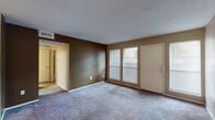 This single-floor residence offers a functional and straightforward layout across several distinct spaces. The living area is generously sized, featuring neutral-toned walls with a darker accent wall, plush gray carpeting, and a textured ceiling. Two windows along the exterior wall allow natural light to fill the room, and a hallway connects the living space to the rest of the unit.

A secondary carpeted room serves as additional living or dining space, featuring warm beige walls and a pass-through opening that connects to the kitchen, enhancing the flow between areas. The galley-style kitchen is equipped with wood-toned cabinetry, dark granite-style countertops, a black refrigerator, dishwasher, and range, all set atop wood-look flooring.

The bedroom is a comfortable, carpeted space featuring taupe walls, a window with natural views, and two separate walk-in closets, providing ample storage. The bathroom includes wood-look flooring, a white tiled tub-shower combo, a toilet, and a vanity with dark countertops and a large mirror, completing the practical design of this well-organized unit.